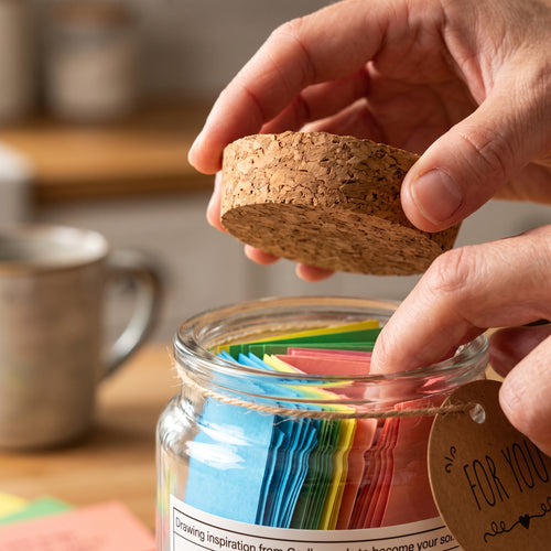 A hand lifting the cork lid off the Bible verse jar in a sunlit kitchen